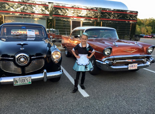 A young girl in a retro outfit stands between two classic cars, a black one and a red one, in a parking lot.