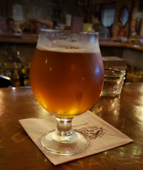 A glass of amber beer sits on a wooden bar top, with a blurred background of patrons and bar items.