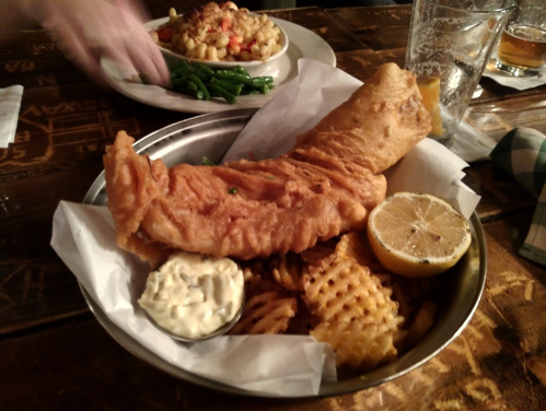 A plate with a large piece of fried fish, waffle fries, tartar sauce, and a lemon wedge on a wooden table.
