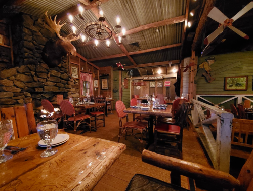 Cozy restaurant interior with wooden tables, stone walls, and rustic decor, featuring a moose head and warm lighting.