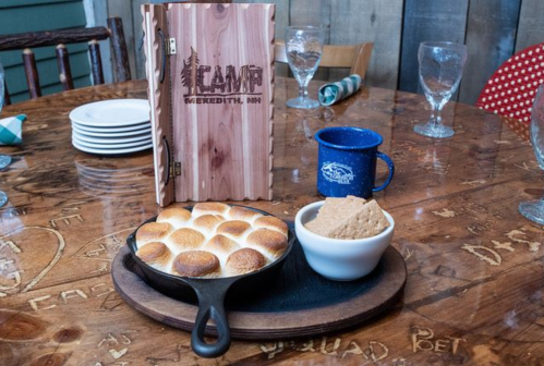 A wooden table with a skillet of baked goods, a bowl of cookies, a blue mug, and a wooden menu at a rustic setting.