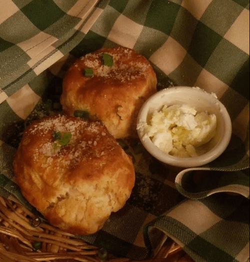 Two golden biscuits topped with green onions, served in a basket with a small dish of creamy butter.