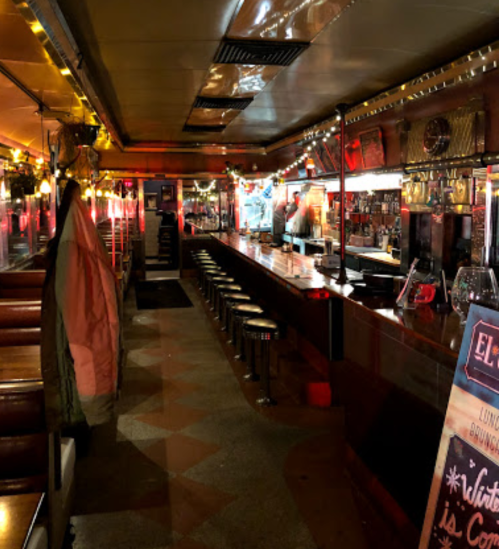 A dimly lit bar interior with a long counter, stools, and festive decorations, creating a cozy atmosphere.