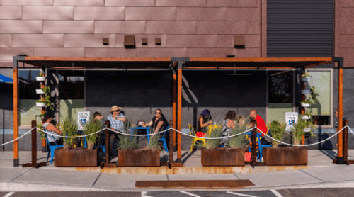 Outdoor dining area with people seated at tables, surrounded by plants and a modern building backdrop.