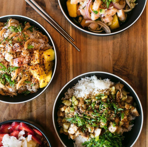 Three bowls of colorful dishes on a wooden table, featuring rice, seafood, mango, and fresh herbs.