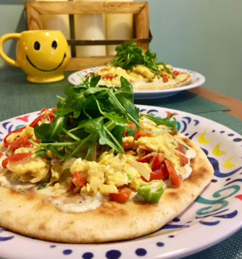 A colorful plate of flatbread topped with scrambled eggs, tomatoes, and arugula, with a cheerful mug in the background.