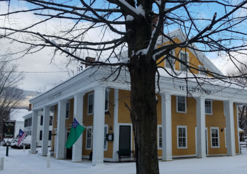 A yellow building with white columns, snow-covered ground, and flags outside, surrounded by bare trees.