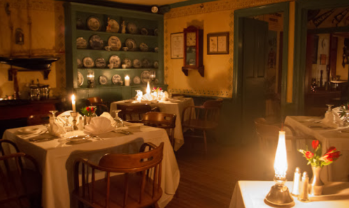 Cozy restaurant interior with candlelit tables, vintage decor, and shelves displaying plates. Warm, inviting atmosphere.