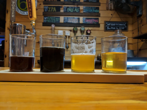 Four glasses of beer in a flight, showcasing different colors and styles, on a wooden bar top.