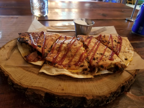 A wooden platter holds sliced quesadillas drizzled with sauce, accompanied by a small cup of dipping sauce.