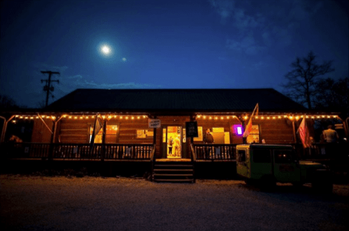 A cozy wooden building illuminated by string lights at night, with a full moon overhead and a green vehicle parked nearby.