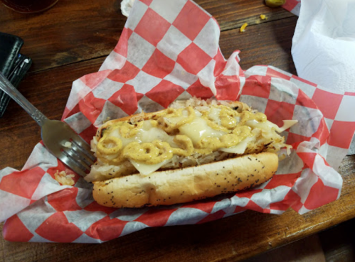 A hot dog topped with melted cheese and mustard, served in a red and white checkered paper on a wooden table.