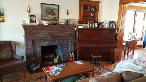 Cozy living room with a brick fireplace, piano, armchair, and wooden shelves filled with photos and decor.