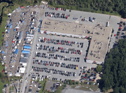 Aerial view of a large parking lot filled with cars, alongside a bustling outdoor market with tents and vendors.