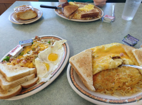 Two breakfast plates with eggs, toast, hash browns, and a cinnamon roll in the background. Condiments on the side.