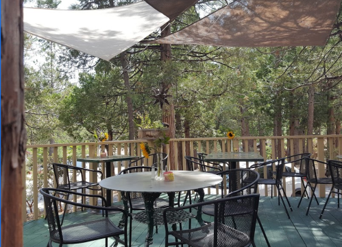 A cozy outdoor seating area with black chairs and tables, shaded by fabric, surrounded by trees.