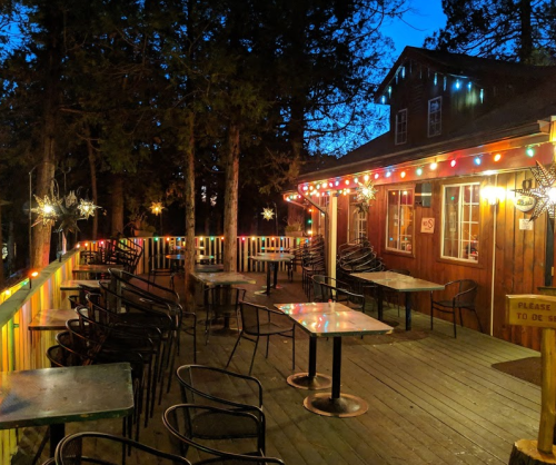 A cozy outdoor dining area with tables and chairs, decorated with colorful lights, surrounded by trees at dusk.