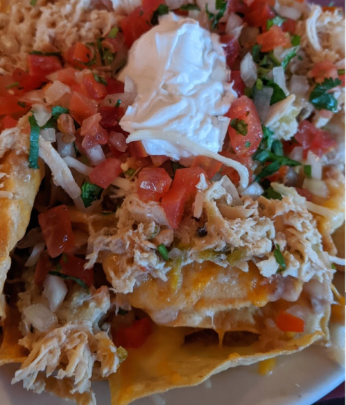 A plate of nachos topped with shredded chicken, diced tomatoes, onions, cilantro, and a dollop of sour cream.