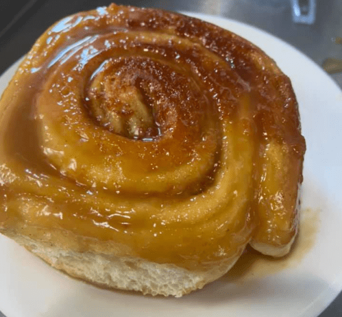 A close-up of a sticky cinnamon roll on a white plate, glistening with caramel glaze.