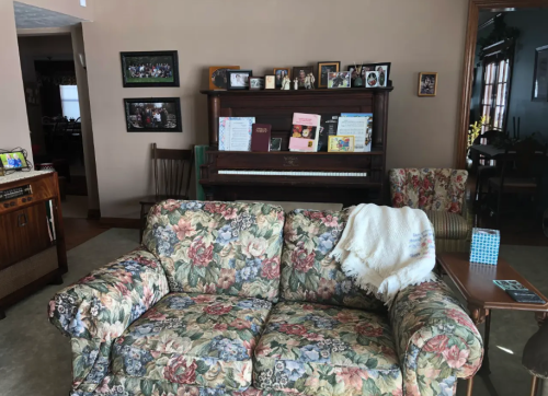A floral-patterned couch in a cozy living room with a piano and family photos on the wall.