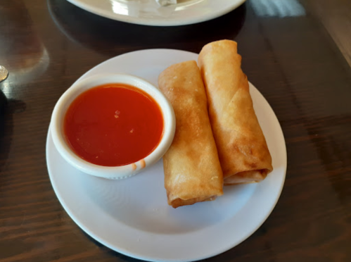 Two crispy spring rolls on a plate with a small bowl of red dipping sauce.