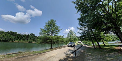 A serene lakeside scene with a car parked near the water, surrounded by trees and a clear blue sky.