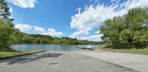 A serene lake view with a wooden dock, surrounded by lush greenery and a bright blue sky with fluffy clouds.