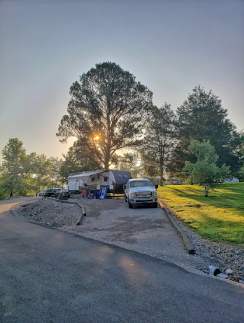 A camper and truck parked under a large tree, with sunlight filtering through the leaves in a serene outdoor setting.