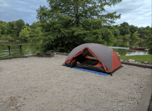 A tent set up on gravel near a calm lake, surrounded by trees and a kayak in the background.
