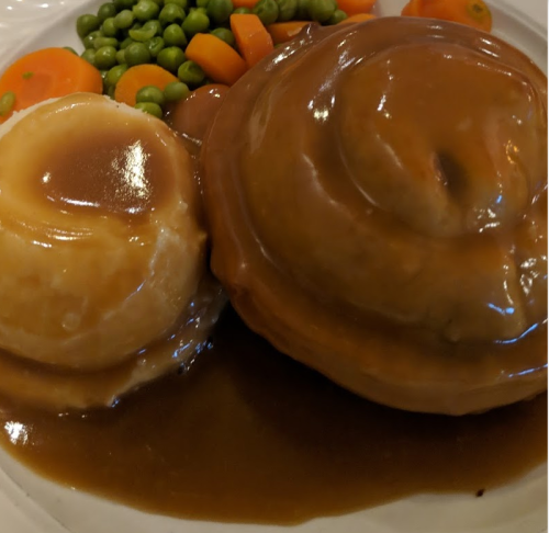A plate of mashed potatoes with gravy, a meat pie, and a side of peas and carrots.