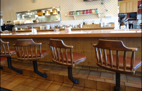 A cozy diner interior featuring a wooden counter with empty chairs and a view of the kitchen area.