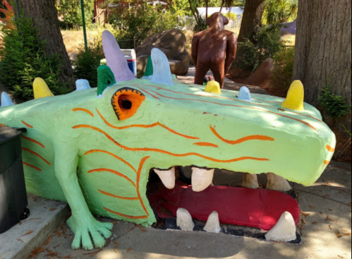 Colorful dragon sculpture with a wide-open mouth, surrounded by trees and other artistic figures in a park setting.