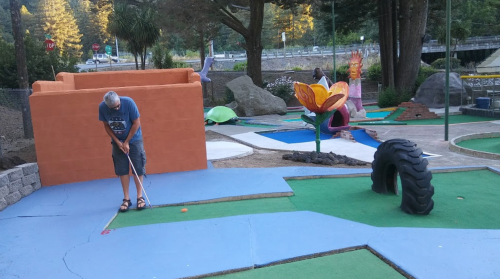 A man putts on a colorful mini-golf course with whimsical decorations and a large flower sculpture in the background.
