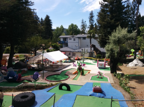 A colorful mini-golf course with various obstacles, surrounded by trees and a building in the background.