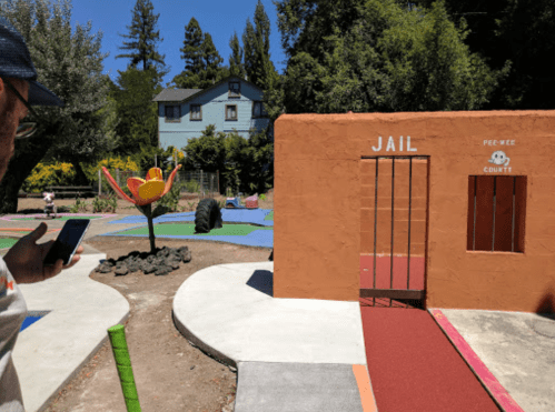 A colorful playground scene featuring a small jail structure and a large flower sculpture, with a person using a phone nearby.