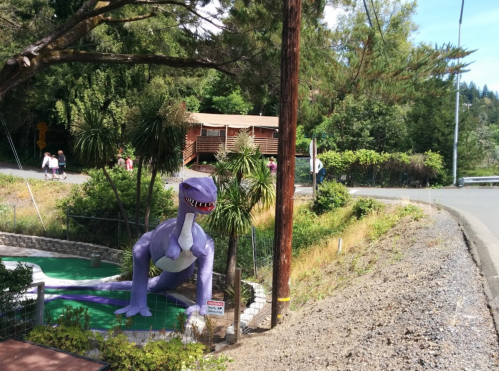 A large purple dinosaur statue stands near a mini-golf course, with trees and a road in the background.