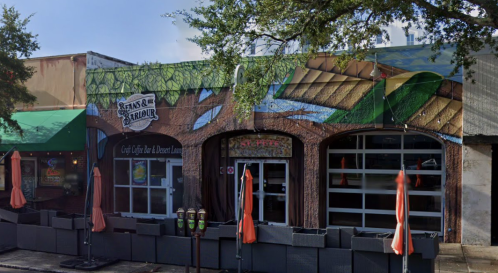 A colorful mural on a brick building with outdoor seating and umbrellas, featuring greenery and a vibrant design.