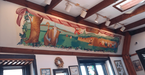 Colorful mural depicting a rural scene with trees, animals, and people working in a field, set in a cozy interior.