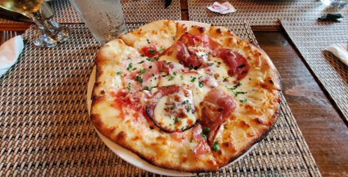 A freshly baked pizza topped with cheese, ham, and herbs, served on a wooden table with drinks in the background.