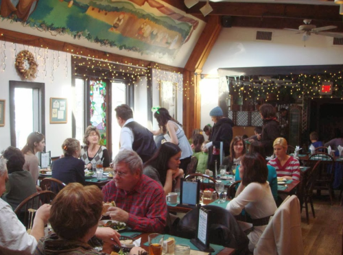 A bustling restaurant interior with diners at tables, a waiter serving, and festive decorations on the walls.