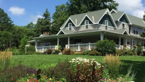 A charming two-story house with a large porch, surrounded by colorful flower gardens and lush green grass.