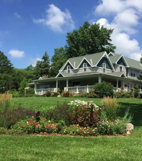 A charming house with a large porch, surrounded by colorful flower beds and lush green grass under a blue sky.