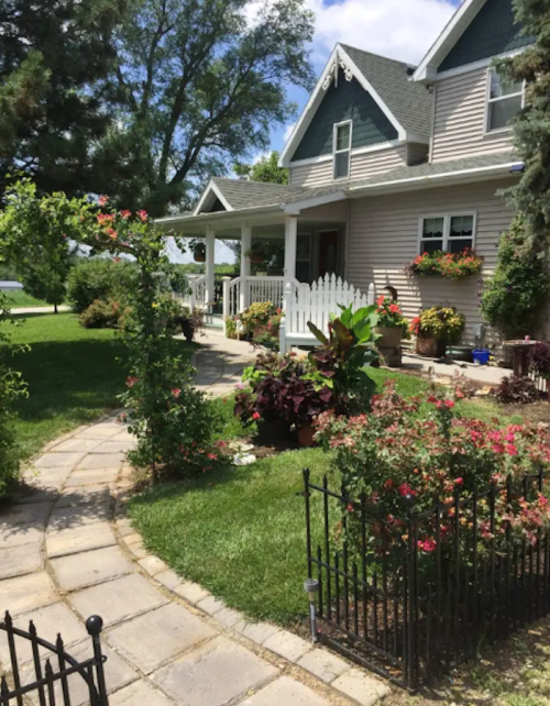 A charming house with a porch, surrounded by a lush garden and colorful flowers, set against a clear blue sky.