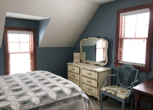 Cozy bedroom with sloped ceiling, a bed, dresser, mirror, and a chair by the window, featuring natural light.