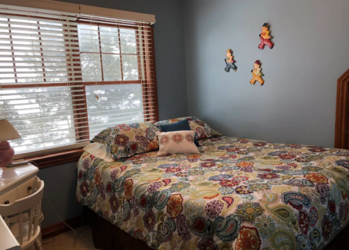 Cozy bedroom with a colorful patterned bedspread, a window with blinds, and decorative wall hangings.