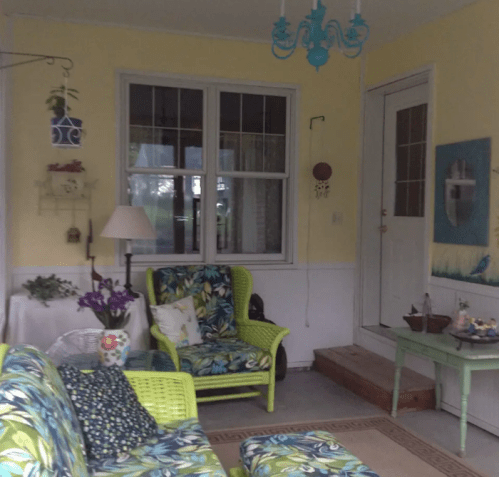 A cozy sunroom with bright green furniture, floral patterns, a chandelier, and yellow walls, featuring a door and decorative art.