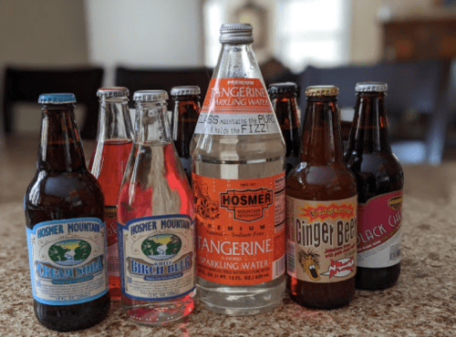 A variety of colorful soda bottles arranged on a countertop, including cream soda, tangerine sparkling water, and ginger beer.