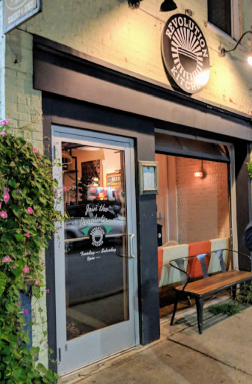 Exterior of Revolution Kitchen, featuring a glass door, colorful bench, and a sign above with a sunburst design.