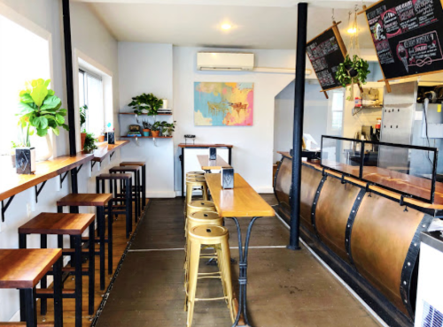 A cozy café interior with wooden tables, metal stools, plants, and a chalkboard menu. Bright and inviting atmosphere.
