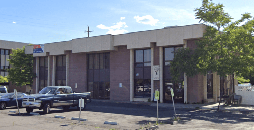 A two-story commercial building with large windows, surrounded by trees and parked cars.